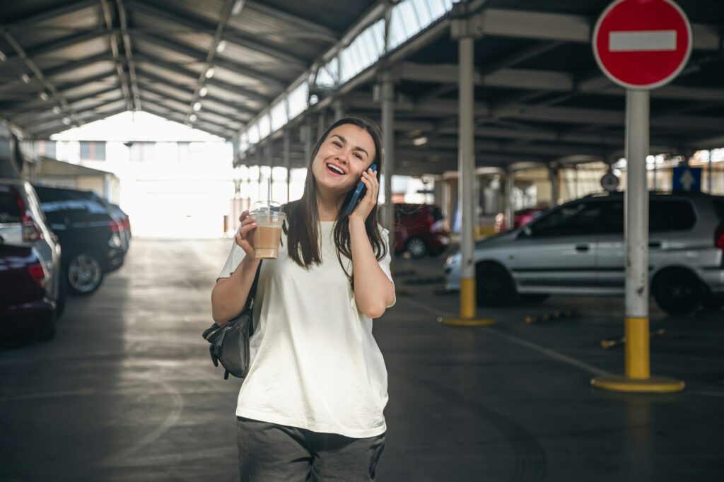 A young woman with coffee talking on the phone in the parking lot.